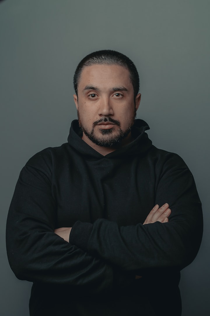 A man with medium-length wavy hair, wearing a teal work jacket, stands looking directly at the camera with a serious expression. The background features blurred white containers, suggesting a workplace environment.